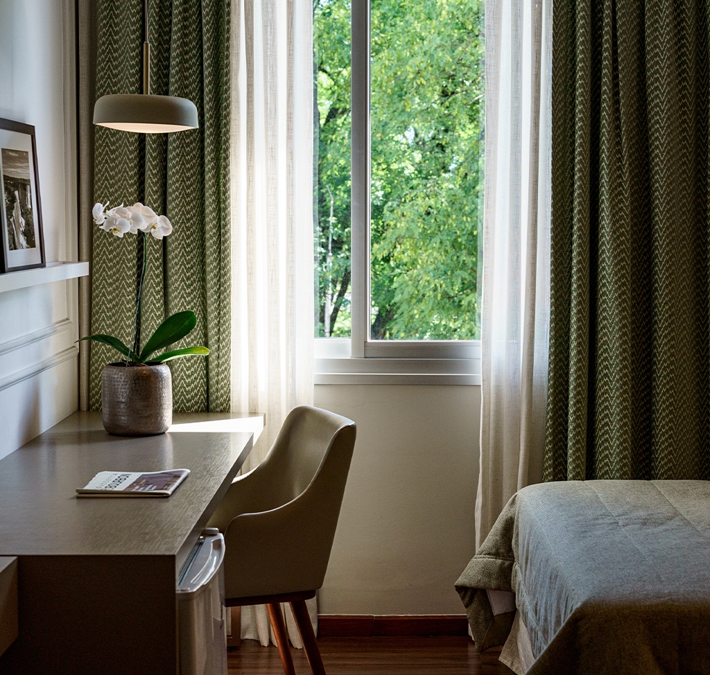 Canto de quarto bem iluminado com escrivaninha de madeira, cadeira estofada, orquídea branca e janela com vista para árvores. Cortinas verdes com estampa em ziguezague e parte de uma cama completam o ambiente.